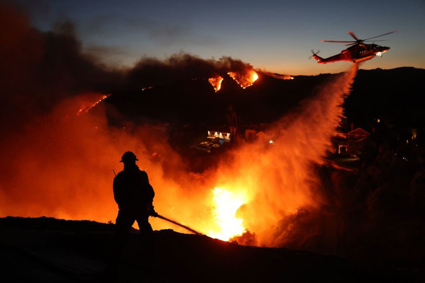 Fire personnel respond to homes destroyed while a helicopter drops water as the Palisades Fire burns on January 7.