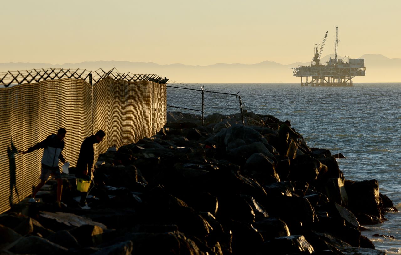 People walk along rocks with offshore oil and gas platform Esther in the distance in Seal Beach, California, on January 5.