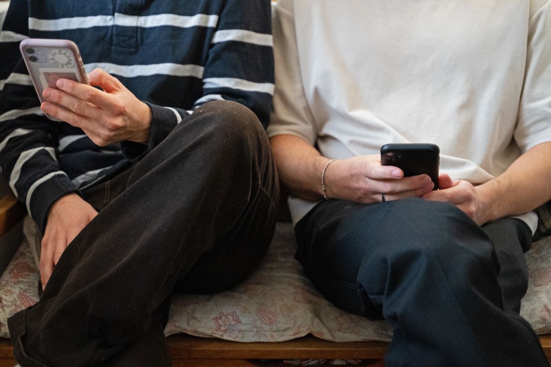 Dos jóvenes sentados uno junto al otro en un banco, con sus teléfonos inteligentes en las manos.