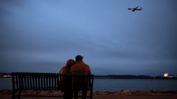 People sit on a bench and watch an airplane fly over the Potomac river during a candlelight vigil held for the victims of the DCA flight collision at Rivergate City Park on February 05, 2025 in Alexandria, Virginia. An American Airlines flight from Wichita, Kansas collided midair with a military Black Hawk helicopter while on approach to Ronald Reagan Washington National Airport on January 29, 2025 outside of Washington, DC. According to reports, there were no survivors among the 67 people onboard both aircraft.