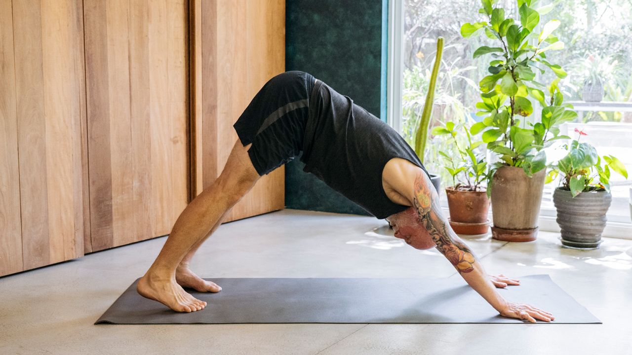 Flexible man in his 60s with bald head wearing shorts and vest bending forwards on yoga mat, in downward facing dog position