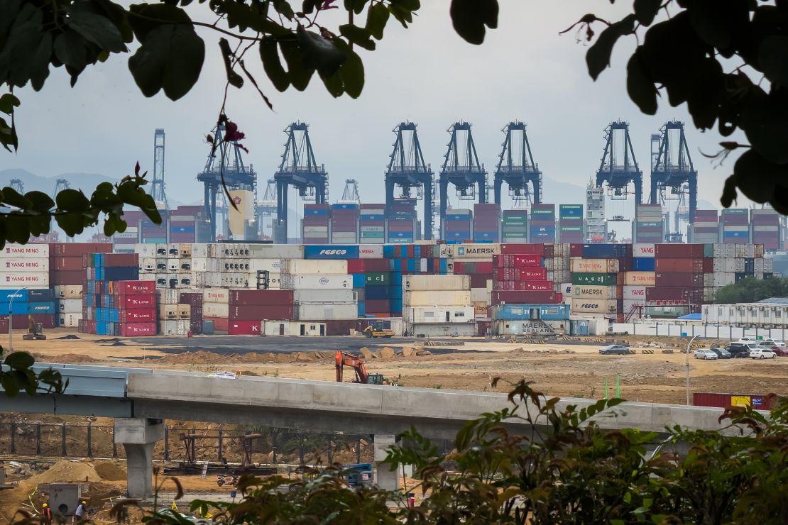 Stacked shipping containers, gantry cranes, and a construction site are seen at the Yantian International Container Terminal through tree branches, with workers and machinery visible in the foreground, on April 12, 2025 in Shenzhen, China.