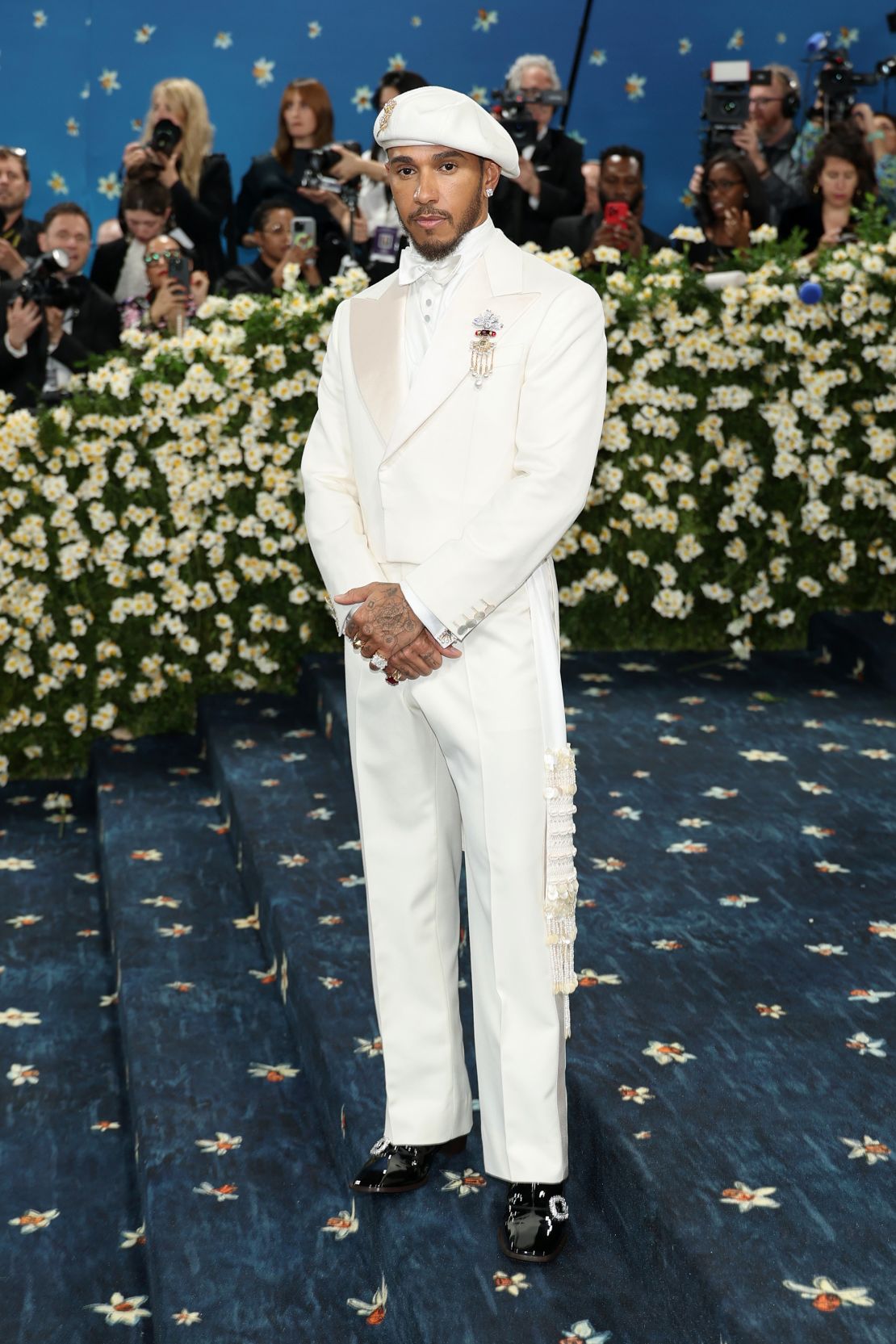 Met Gala co-chair Lewis Hamilton stuns in a custom white Wales Bonner tuxedo. “The color ivory denotes purity and status,” and “ancestral beads and freshwater pearls shine with garnet-colored diamonds… eternal amulets,” stylist Eric McNeal wrote on Instagram.