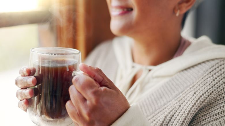 Home, hands and old woman with coffee, window view and morning routine with retirement. Apartment, mature person and senior lady in lounge, espresso and resting with break, smiling and closeup