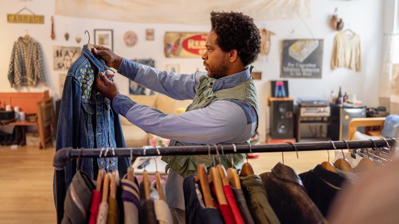 Young man arranging clothes on hangers in a vintage second-hand shop, creating an inviting display of unique garments for customers