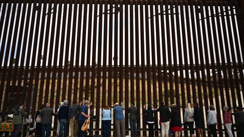 People lay their hands on the border wall and pray towards Mexico during Pastor Seth Clark's last service for The Border Church at the stretch of the US-Mexico border fence known as "Whiskey 8" in San Diego, California on May 25, 2025. As pastor Seth Clark retires from his leadership role with The Border Church the mission of the interdenominational Christian ministry to maintain presence and sacramental solidarity appears more challenging now than ever. While weekly binational communion services started in 2011 on both sides of the US and Mexico border, the US side of the International Friendship Park has remained closed since February 2020 with fears it will never reopen. The recent addition of concertina wire along the top and bottom of the border wall bollards features sharp barbs akin to a crown of thorns meant to deter migrant crossings. (Photo by Patrick T. Fallon / AFP) (Photo by PATRICK T. FALLON/AFP via Getty Images)          