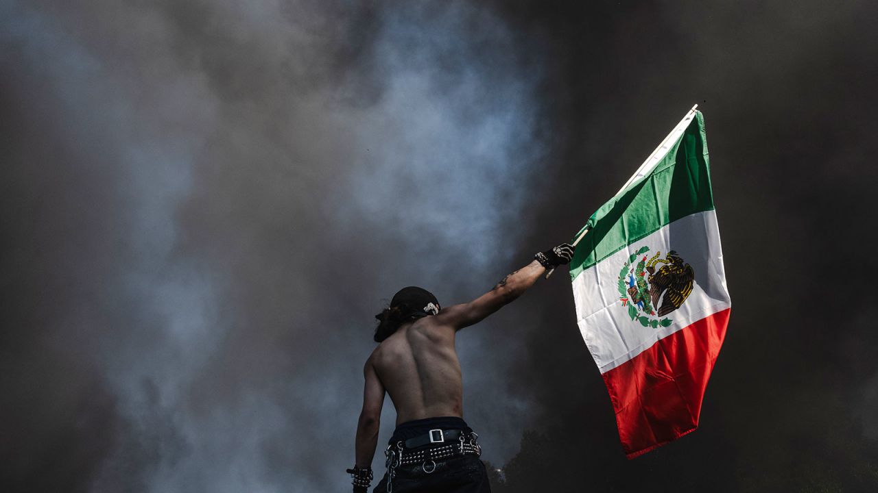 A protester holds up a Mexican flag while standing in front of smoke billowing from burning cars on June 8, 2025 in Downtown Los Angeles, California.