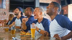 Multi Cultural Group Of Male Friends Wearing Team Colores Watching Game In Sports Bar Eating Food