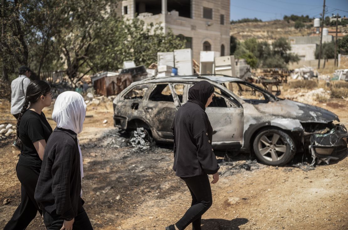 Palestinian women walk past a charred vehicle, as they survey the destruction wrought Israeli settlers in the West Bank village of Kafr Malik, on Thursday.