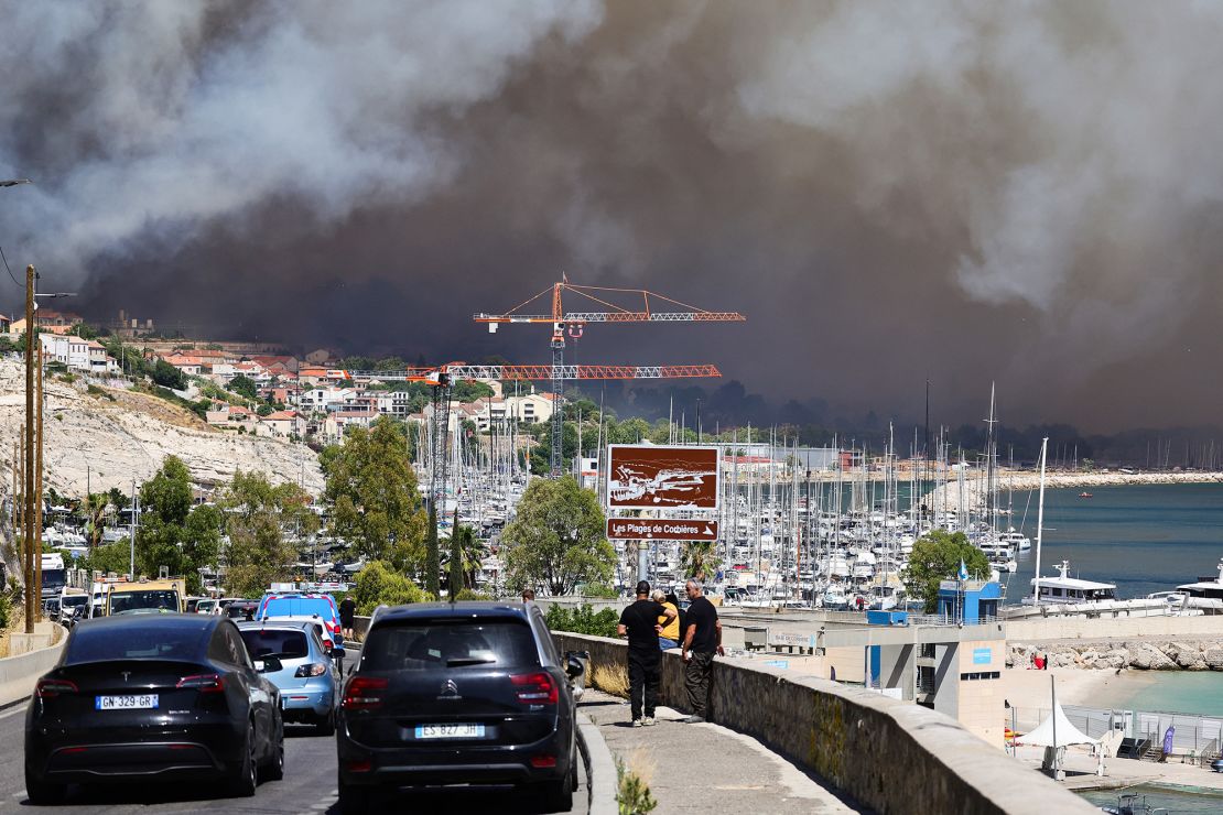 People look on near the Plage des Corbieres, in Marseille, southern France on July 8, 2025, as a smoke from a wildfire rages in the background.