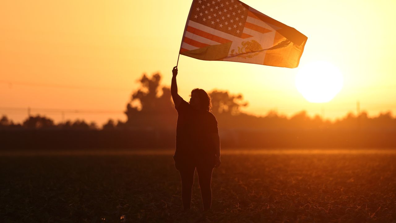 CAMARILLO, CALIFORNIA - JULY 10: Community member Ana waves an American-Mexican flag in a show of support for detainees near federal agents blocking protestors during an ICE immigration raid at a nearby licensed cannabis farm on July 10, 2025 near Camarillo, California. Ana said she knows some of those detained and their families. Protestors stood off with federal agents for hours outside the farm in the farmworker community in Ventura County. A Los Angeles federal judge is set to rule Friday on a temporary restraining order which would restrict area immigration enforcement operations. (Photo by Mario Tama/Getty Images)