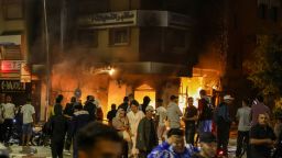 TOPSHOT - People pass by a bank building set on fire during a youth-led demonstration demanding reforms in the healthcare and education sectors in Sale on October 1, 2025. Two people were killed when officers opened fire on a group of people attempting to "storm" a police station in Morocco on October 1, state media said, as protests -- sometimes violent -- roil the north African nation. Demonstrations have convulsed Morocco for several days, urged on by the GenZ 212 group, a recently formed collective based on the Discord web platform whose organisers remain unknown. (Photo by Abdel Majid BZIOUAT / AFP) (Photo by ABDEL MAJID BZIOUAT/AFP via Getty Images)          