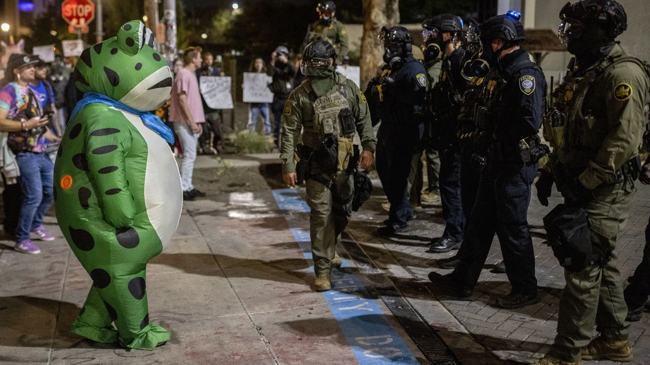 A protester in a frog costume stands in front of a line of federal law enforcement officers outside an ICE facility in Portland, Oregon.