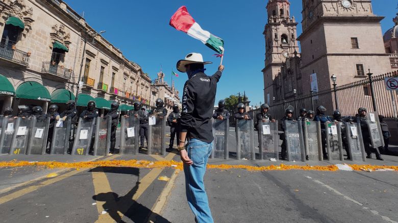 A protester waves a Mexican flag in front of police officers during a demonstration against the assassination of Uruapan's mayor at the Government Palace in Morelia, Michoacan state, Mexico, on November 3, 2025. The Mexican government reported on November 2 that the mayor of Uruapan, Carlos Manzo, who was killed the previous night during a public event in the western state of Michoacan, had been under official protection since December last year. (Photo by Jordi LEBRIJA / AFP) (Photo by JORDI LEBRIJA/AFP via Getty Images)          