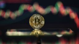 An illustration photograph taken on November 22, 2025, shows a gold plated souvenir Bitcoin coin reflected in a mirror and arranged for a photograph in front of a computer screen displaying the Bitcoin monthly price chart in a residential property in Guildford, south of London. The value of bitcoin has fallen sharply since reaching record heights last month, above $126,000 at the start of October.