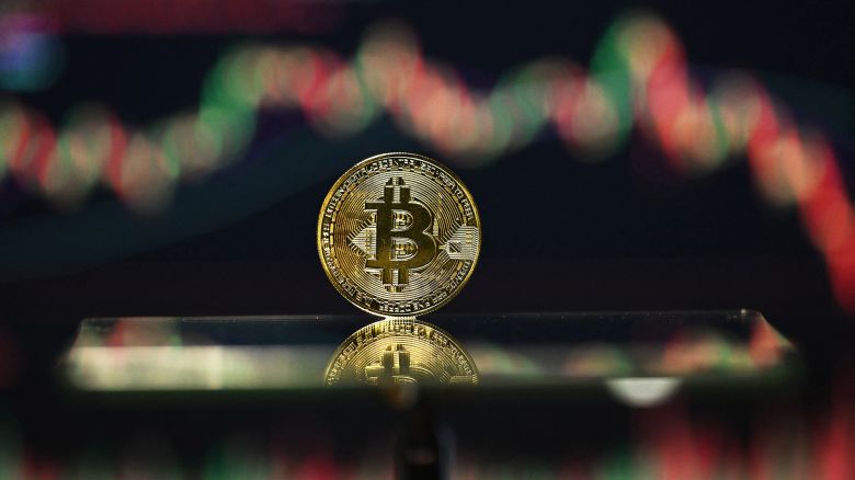 An illustration photograph taken on November 22, 2025, shows a gold plated souvenir Bitcoin coin reflected in a mirror and arranged for a photograph in front of a computer screen displaying the Bitcoin monthly price chart in a residential property in Guildford, south of London. The value of bitcoin has fallen sharply since reaching record heights last month, above $126,000 at the start of October.