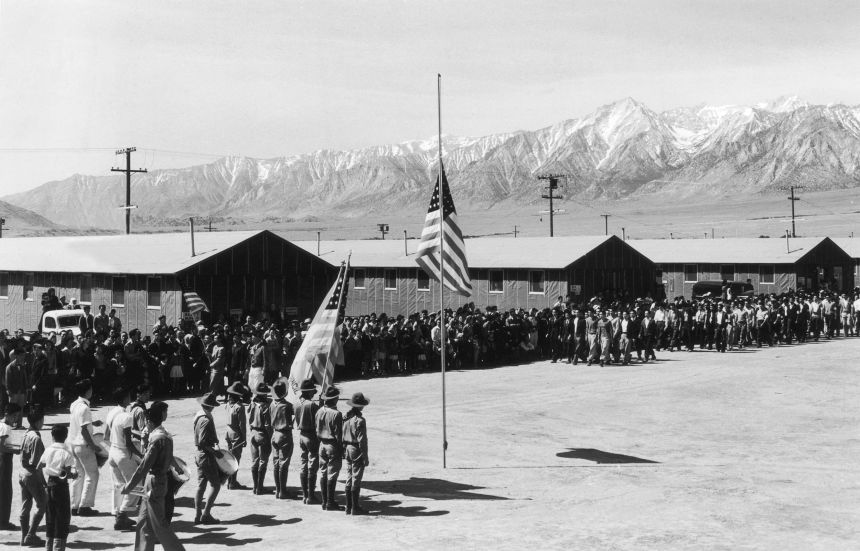 Soldiers, a small marching band and civilians lined up near a flagpole outside barracks at Manzanar Japanese-American internment camp in 1943 near Independence, California, during World War II. Mount Whitney, the highest peak in the United States, is visible in the background.