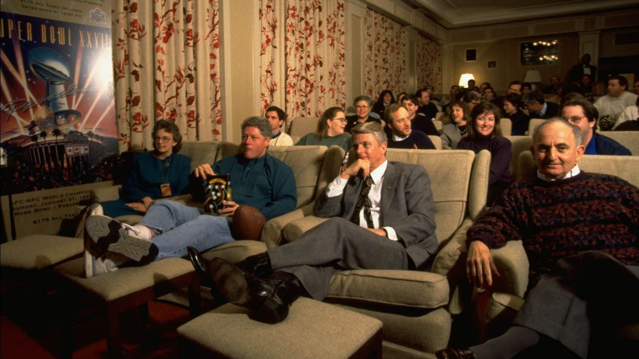 President Bill Clinton (C) watching Super Bowl in family theater with governor guests and White House staffers, munching popcorn, with sneakered feet on ottoman and a football at his side in 1994.