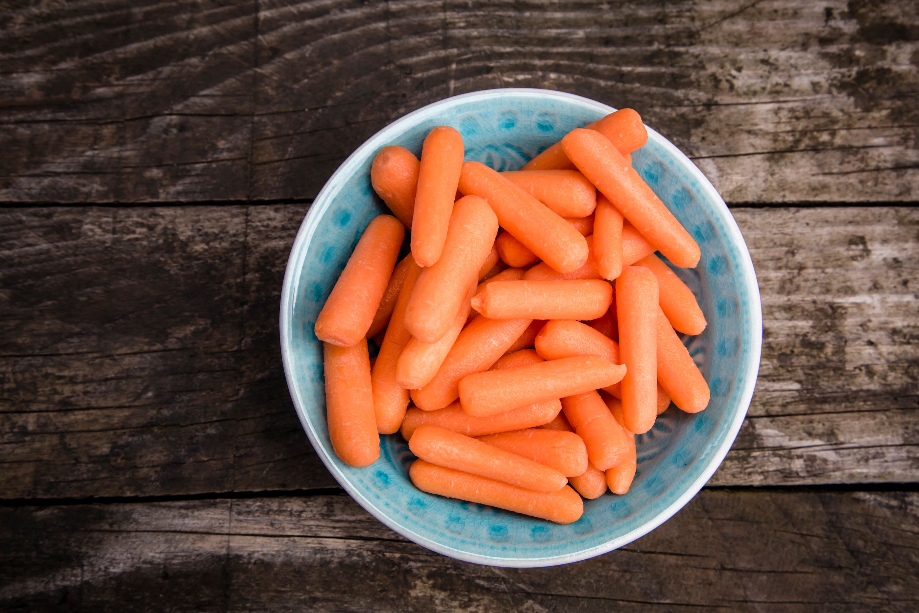 Top-down view of a turquoise bowl overflowing with bright orange baby carrots on a rustic wooden surface, evoking healthy snacking.