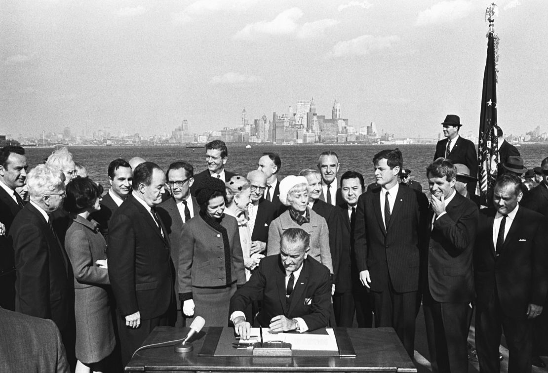 President Lyndon B. Johnson signs the Immigration Bill of 1965 on Liberty Island in New York Harbor with a view of the New York City skyline in the background. Next to the president on his right are First Lady Lady Bird Johnson and Vice President Hubert Humphrey. To the president's left are Senator Edward Kennedy (third from right) and Robert Kennedy (second from right).
