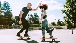 A fun, playful Hispanic Dad and his son play together at a skate park. The boy has a helmet and padding, and is having fun with his father. They give each other a high five.  A depiction of a loving supportive dad.