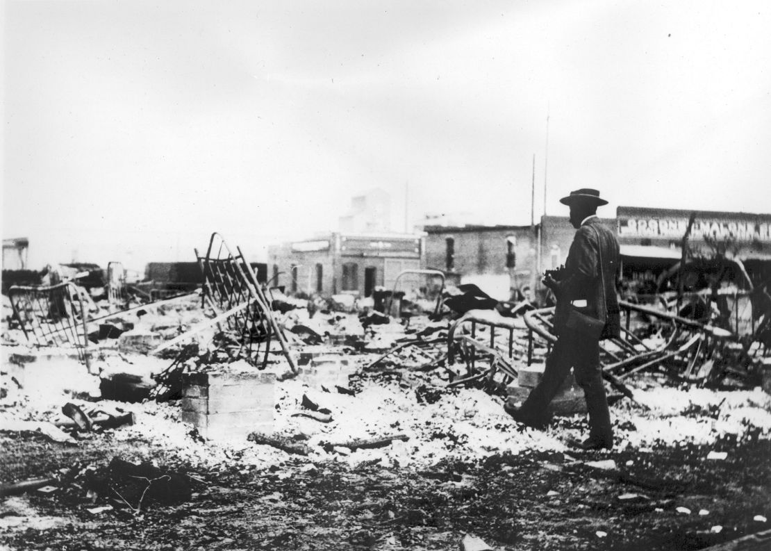 An African American man with a camera looking at mangled bed frames, which rise above the ashes of a burned-out block after the Tulsa Race Massacre, Tulsa, Oklahoma, June 1921.