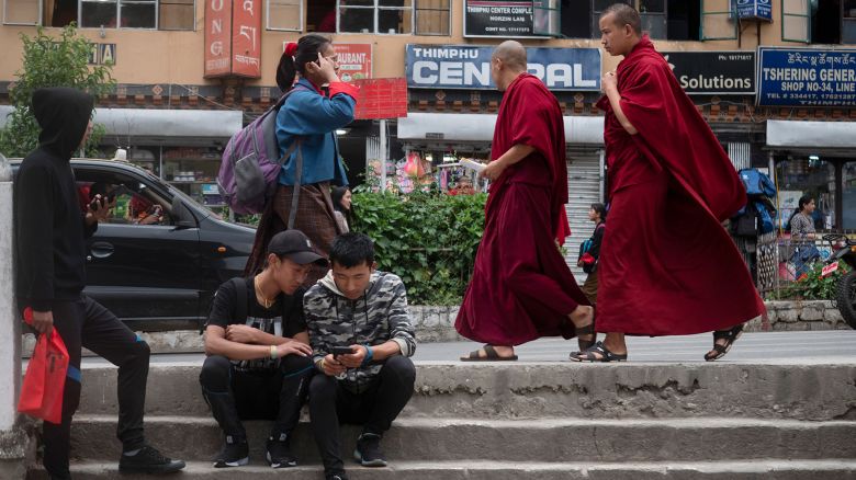THIMPHU, BHUTAN - JUNE14 : Bhutanese teens are seen hanging out using mobile phones as monks stroll by on June 14,2018, in Thimphu, Bhutan. While many are expected to wear the "Gho" which is the traditional and national dress in Bhutan, the younger generation tends to wear western clothes when they are off work or after school. Bhutan is no ordinary place, it is known an open democratic society. Bhutanese society is free of class or a caste system, the country has a very young population: more than half of the population is below the age of 25 years. Perhaps the last great Himalayan kingdom, where a traditional Buddhist culture carefully embraces global developments. The youth in Bhutan today live in a world very different from that known by their parents. Globalization, urbanization and new modes of mass and interpersonal communication have rapidly and radically changed the way young people interact with each other, with their families and with society as a whole. (Photo by Paula Bronstein/Getty Images for Lumix)