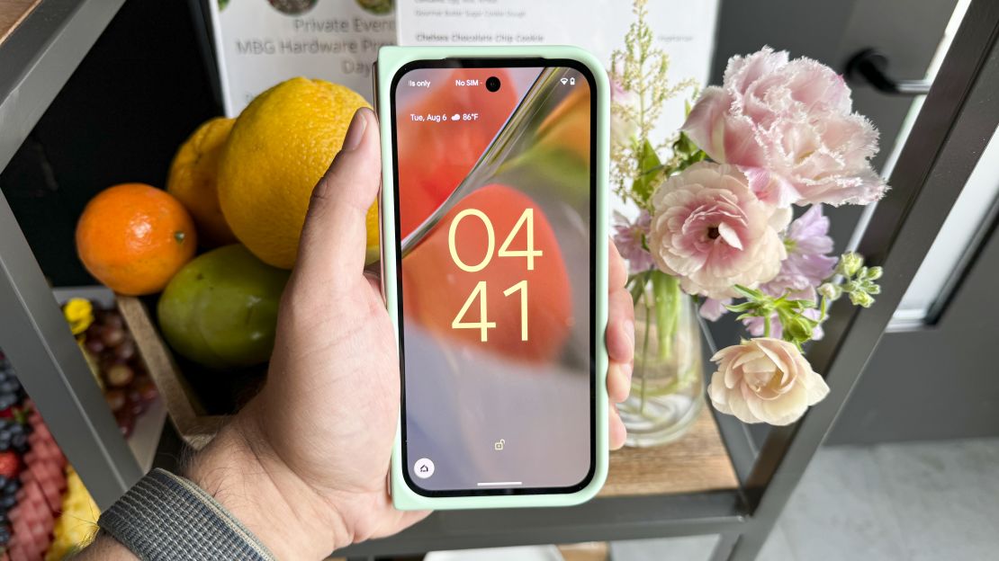 The Google Pixel 9 Pro Fold is in a minty case and closed shut with its display on to the time, in front of flowers.