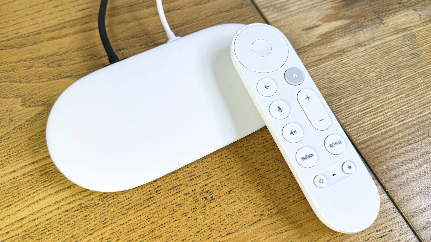 The Google TV Streamer is plugged into two cords, on a wooden surface, with its remote on top of it.