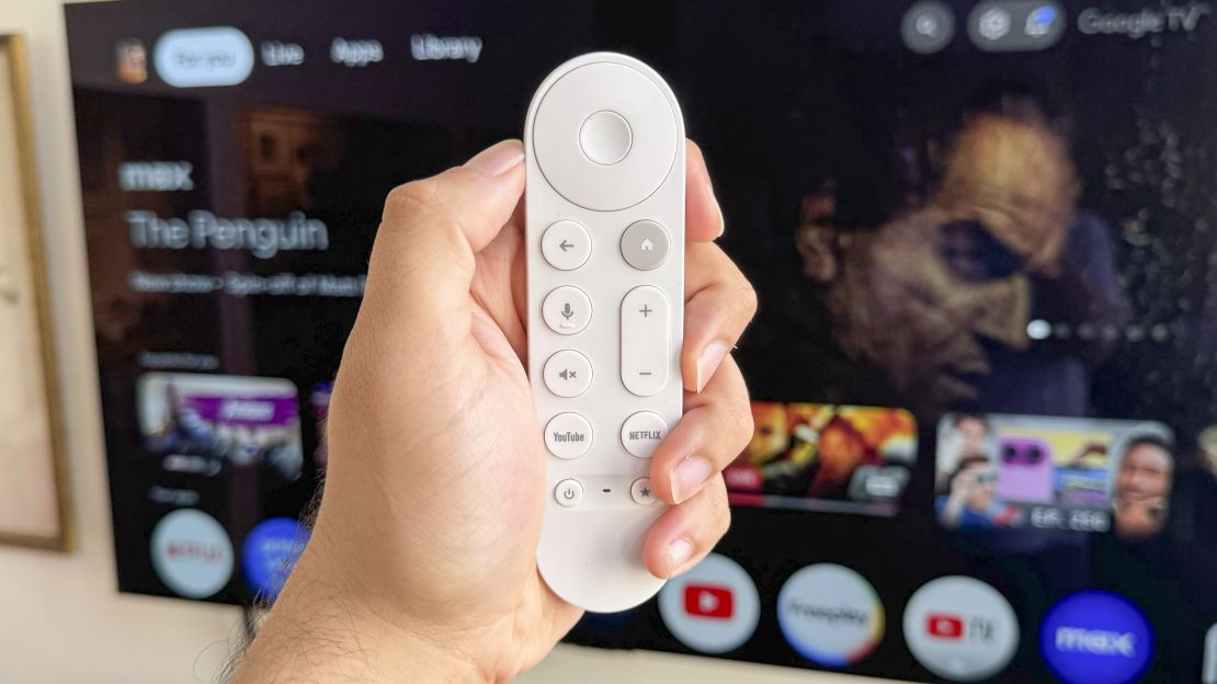 A hand holds the Google TV Streamer remote in front of a TV showing the Google TV home screen, connected to the (not shown) Google TV Streamer