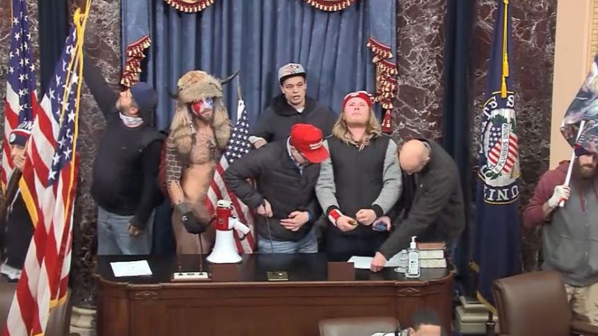 In this photograph included in a statement of facts document by the US District Court for the District of Columbia, Christopher Moynihan, seen at center in the gray hat, stands in the Senate Chamber during the January 6, 2021, attack on the US Capitol. Moynihan has been arrested after allegedly making threats against Democratic House Minority Leader Hakeem Jeffries.
