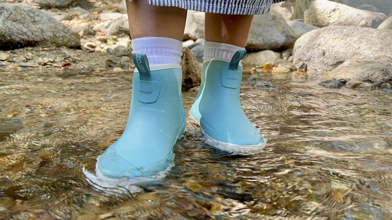 Close up of person in rain boots standing in shallow river.