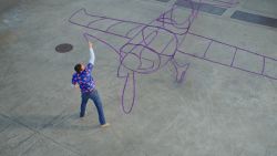 An overhead shot of "Harold" (Zachary Levi), in a purple shirt with red flowers on it and blue jeans, uses his purple crayon to outline a large airplane.