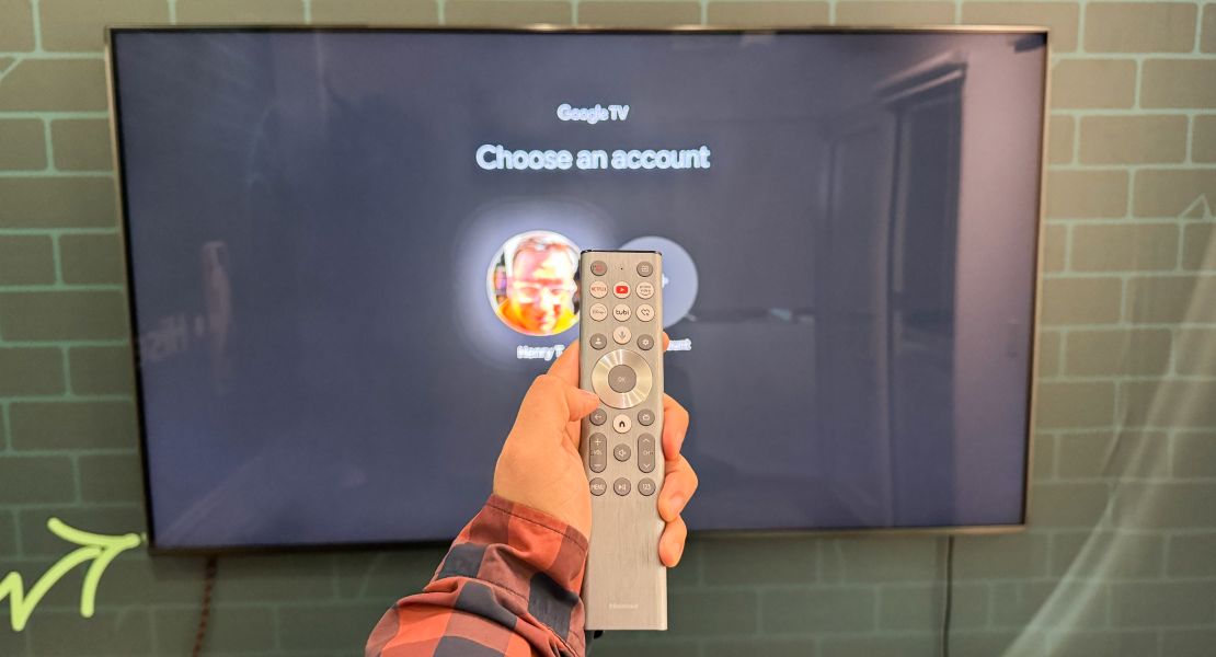 A hand holds the Hisense U9N’s remote in front of the TV, open to its Google TV login menu.