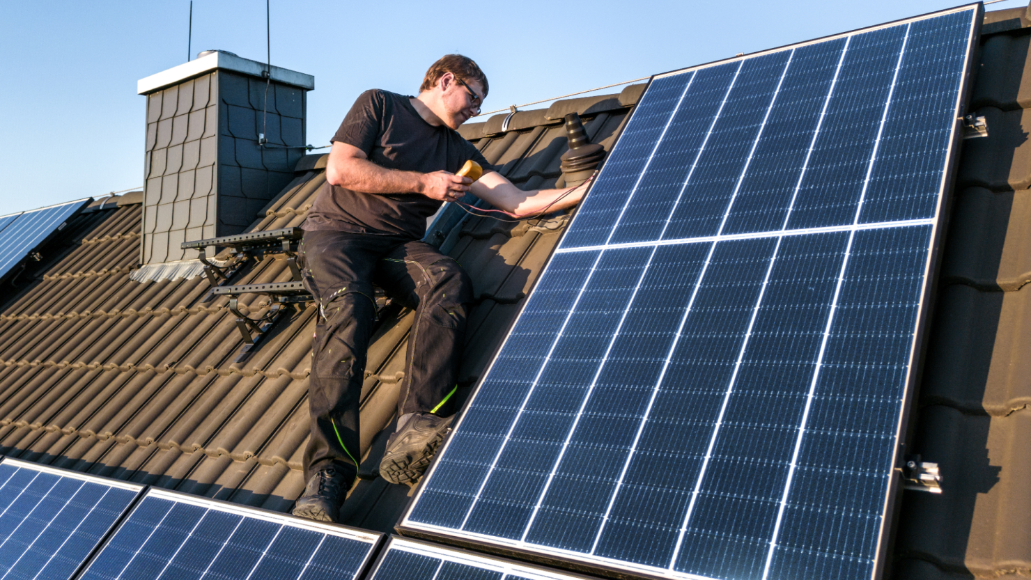 A man sits on a house's roof using a multimeter to check the photovoltaic system.