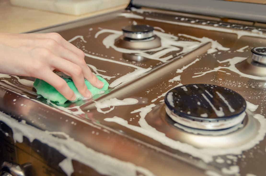 woman's hand washes a gray stainless steel gas stove with a green foam sponge. House cleaning