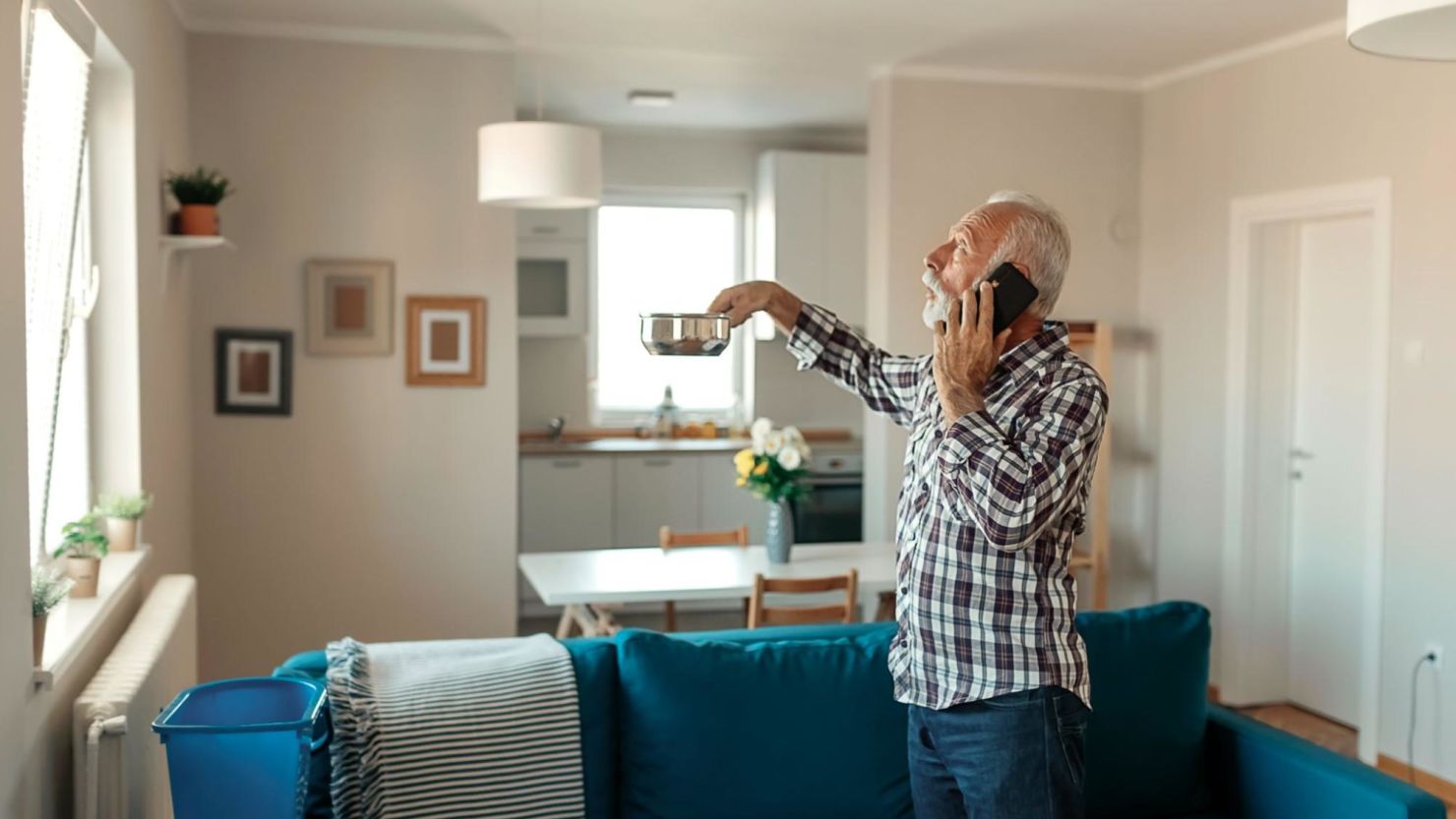 Homeowner calls his home warranty provider as he holds a pot below a leak to catch water.