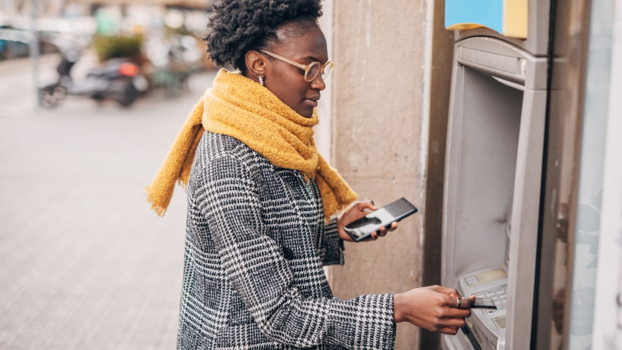 A person using an ATM while holding a cell phone and an ATM card.