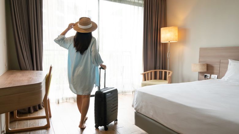 A photo of a person wearing a straw hat walking into a hotel room while pulling a small suitcase