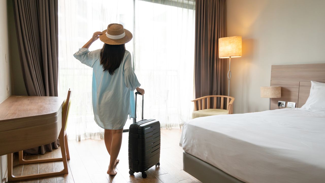 A photo of a person wearing a straw hat walking into a hotel room while pulling a small suitcase