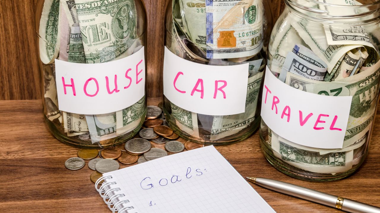 A photo of jars labeled "House", "Car" and "Travel" with cash inside of each jar.