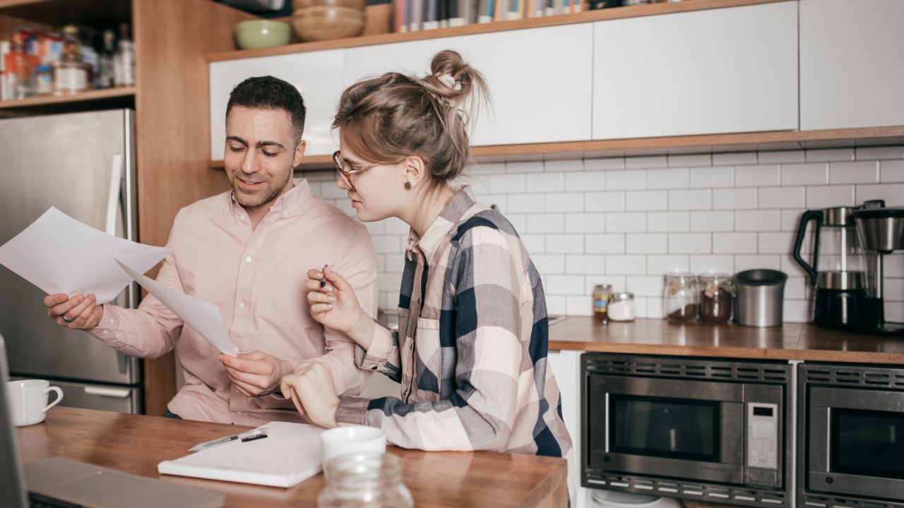A photo of two people talking in a residential kitchen while looking at pieces of paper.