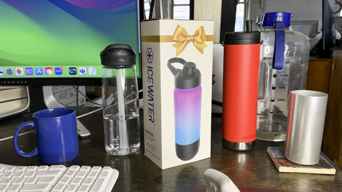 (left to right) On a desk there's a mug, a clear water bottle, the Icewater smart water bottle box , a red thermos, a giant water bottle and a glass