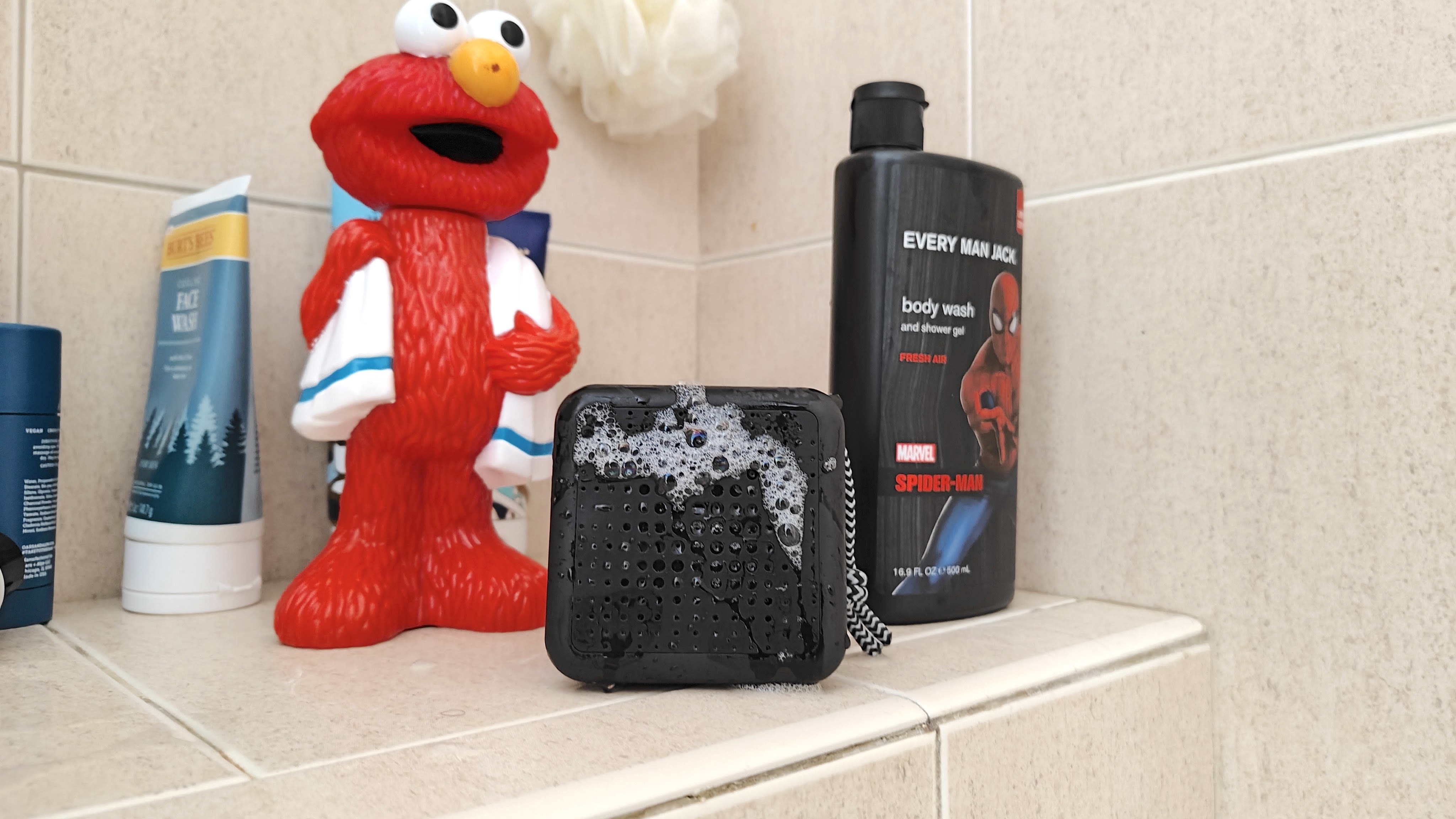 A waterproof speaker and various shampoo bottles, one of which is shaped like Elmo, in a shower.