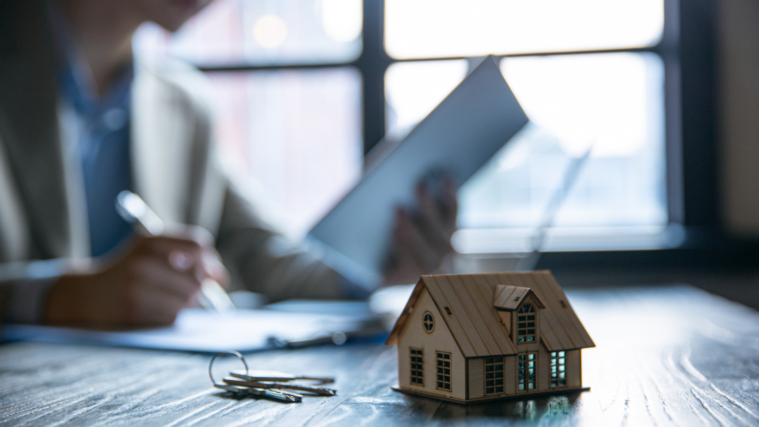 A small model home in the foreground of a homeowner completing documents