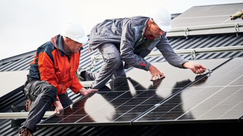 Two solar installers place a solar panel on a roof.