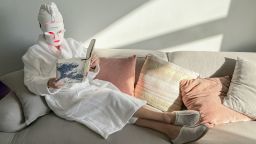 Person relaxing on couch wearing a cozy robe and LED light therapy face mask while reading a book.