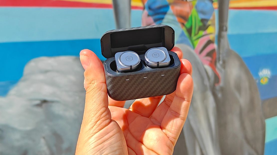 A hand holds encased wireless earbuds in front of a mural.