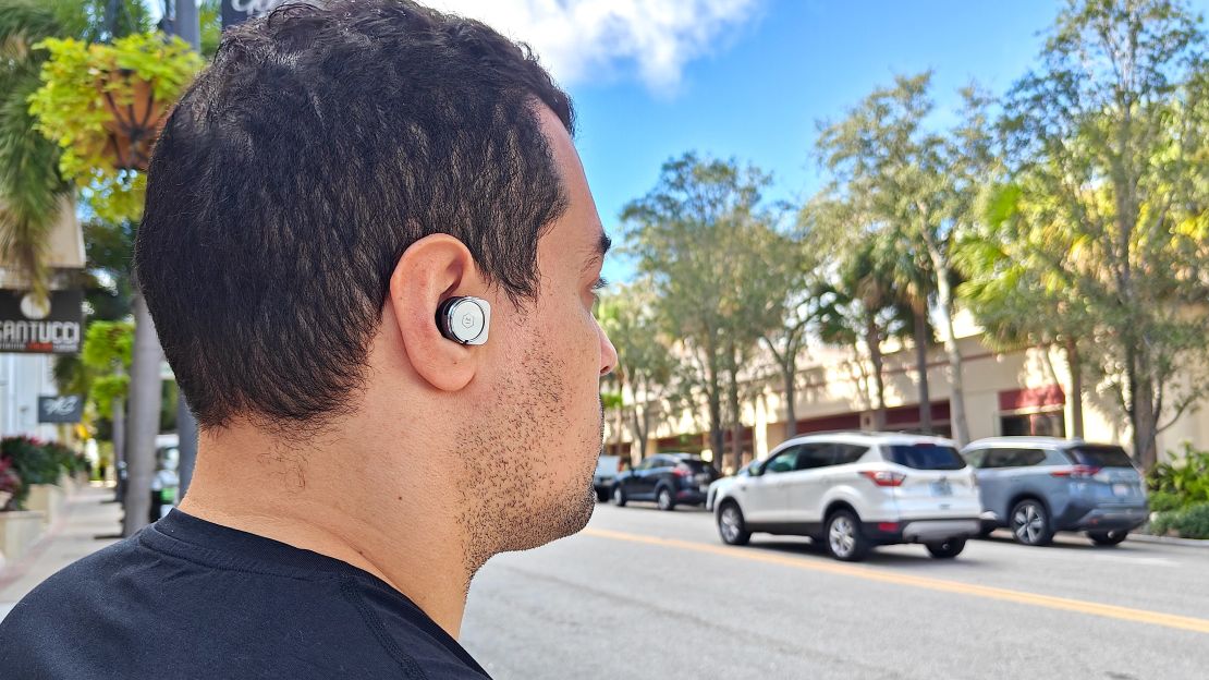 The side profile of a person wearing a silver wireless earbud. He's looking at cars in the street.