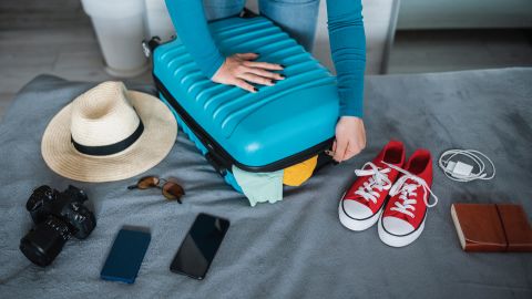 A photo of a person closing a carry-on suitcase on a bed with various items scattered around